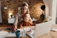 Mid-adult-working-mother-taking-notes-during-conference-call-home